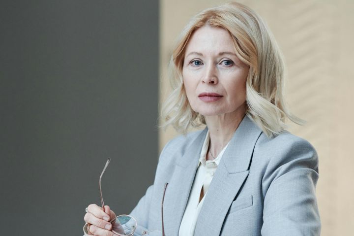 Senior caucasian woman in gray blazer holds papers in a modern workplace setting.