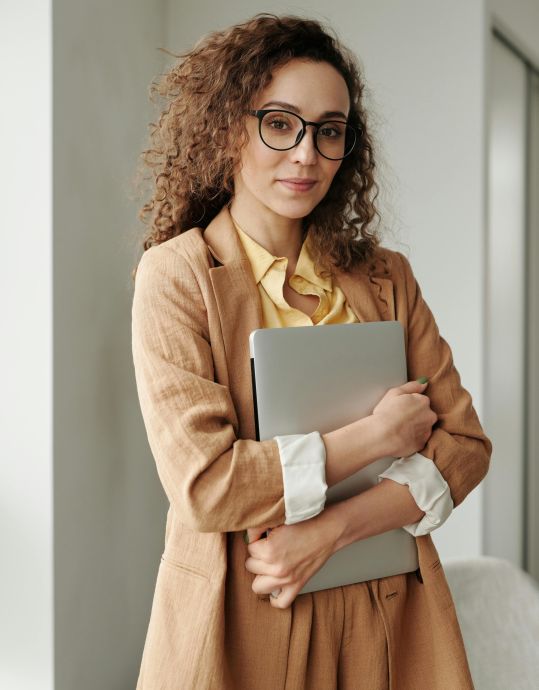 A confident woman in formal attire holding a laptop, standing in an office environment and looking at the camera.